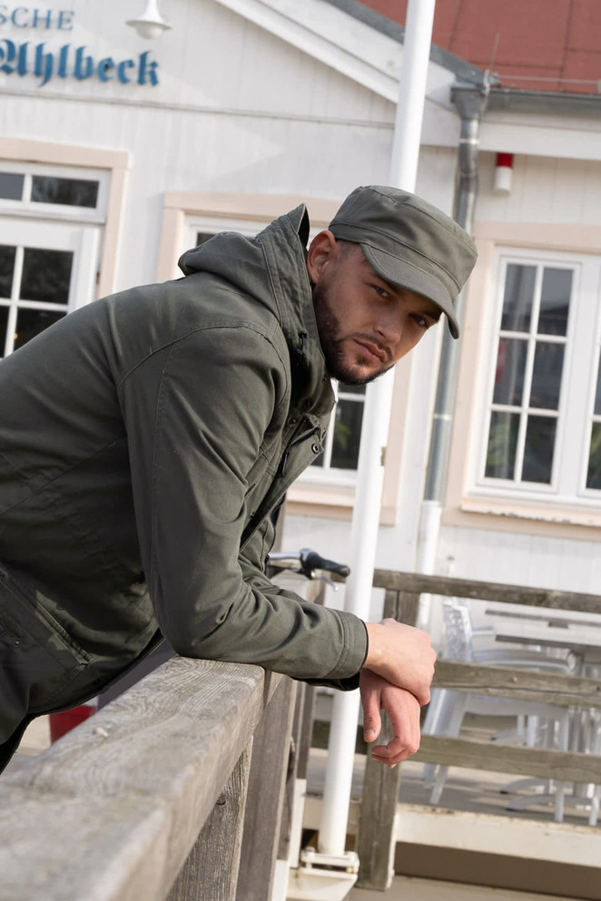 A man leans against a railing wearing an olive green Sports Twill Military Cap and a dark jacket.