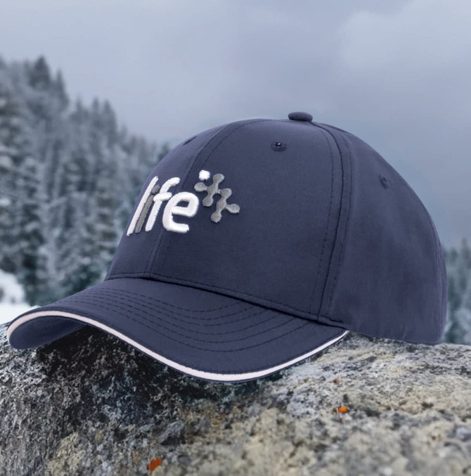 A navy sports cap with sandwich trim, featuring a logo on the front, resting on a stone in a mountainous landscape.
