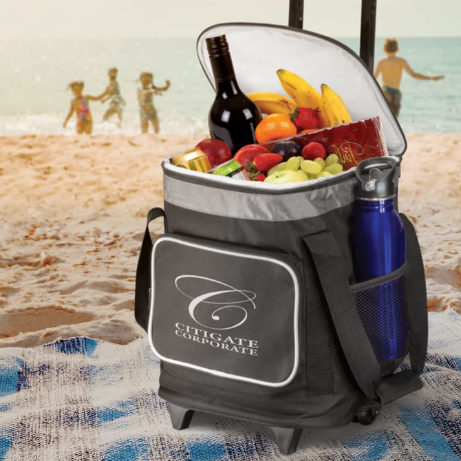 A black cooler trolley with wheels, filled with wine, fruit, snacks, and a blue drink bottle, set on a beach.