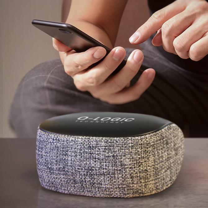 A Cylon Bluetooth speaker in grey fabric with a black top, beside a hand holding a smartphone.