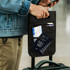 A black Trolley Bag Caddy attached to a suitcase, holding passports, a boarding pass, and a pen.