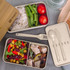 A stackable lunch box in neutral tones, featuring compartments with rice, vegetables, and salad, along with a bamboo lid.