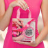A pink gift bag featuring hands and a ribbon design, with a logo on one side, held by a person in a pink outfit.
