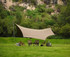 A tan camping canopy with poles, set up in a grassy area surrounded by greenery and rocky terrain.