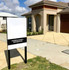 A flat metal sign on a black post stands outside a modern beige house on a paved driveway.