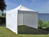 A white marquee tent stands on grass near a lake, featuring a peaked roof and open sides.