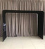 A black fabric arch frame with a tension design, set against a neutral curtain backdrop. Features a logo on one side.