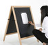 A black magnetic A-frame chalkboard with a wooden frame, being adjusted by a person in a white shirt.