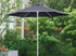 A black café umbrella with a sturdy white pole, set against a lush green garden backdrop.