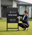 A large black steel lawn sign with directional arrow and text about an open home being arranged by a woman in business attire.