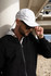 A person wearing a white cap and sunglasses stands near a sandy area, dressed in a black jacket.
