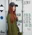 A woman with long brown hair wears a navy cap and an olive-green jacket, posing against a shipping container.