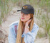 A woman with long blonde hair wears a black premium twill cap, seated on sandy ground with grass in the background.
