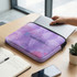 A large purple laptop sleeve with a logo, resting on a table beside a notebook and a plant.