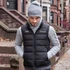 A black unisex puffer vest worn over a grey sweater, paired with a grey beanie. The vest features a logo.
