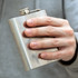 A stainless steel hip flask held in a hand, featuring a screw-top lid and a logo on the front.