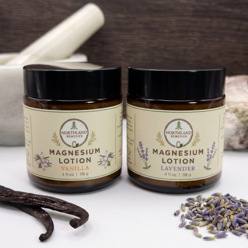 Northland Remedies Lotions in a 4 oz amber glass jar, displayed next to an open jar of thick white topical cream, vanilla beans, lavender buds, and a white ceramic mortar and pestle on a dark wood background.