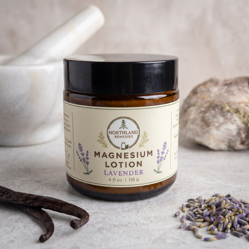 Northland Remedies Lavender Magnesium Lotion in a 4 oz amber glass jar, displayed next to an open jar of thick white topical cream, vanilla beans, lavender buds, and a white ceramic mortar and pestle on a dark wood background.
