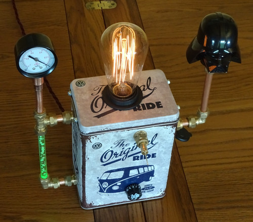 A tin, with the image of a VW camper wagon, has been converted to a unique hand-built steampunk dimmable lamp, with manometer and illuminated USB charging port. 
This artwork has a rotary ‘skull and crossbones’ switch/dimmer and an Edison-style E27 bulb to brighten the corners of your bothy. It has been fitted with rubber feet (not your bothy). The product is about 26cm wide, 35 cm high and 15 cm deep. 
On the left side green bubbles flicker, above which resides an upcycled manometer (pressure gauge) has a scale of 30 lb/sq in. Normally this will read zero. 
To its right it has a green illuminated USB charger port, itself powered by a 30-watt stabilised power supply. Rubber feet have been added to the base.
The 10mm copper piping has brass fittings that are compression type with copper olives (not the type in a Martini, shaken not stirred).
On the right-hand side is an upcycled candy container Darth Vader. 
The burgundy fabric double insulated 3 core 1.6 metre cable includes an in-line black traditional Torpedo 6-amp rocker switch, which disconnects both the Live and Neutral, and complies with BS EN 6108-2-1.  This cable is terminated with a black 13-amp plug, which complies with BS 1363.  The other end is terminated with a black IP68 compression TRS M12 cable stuffing gland. Really, we didn’t just make that up.
