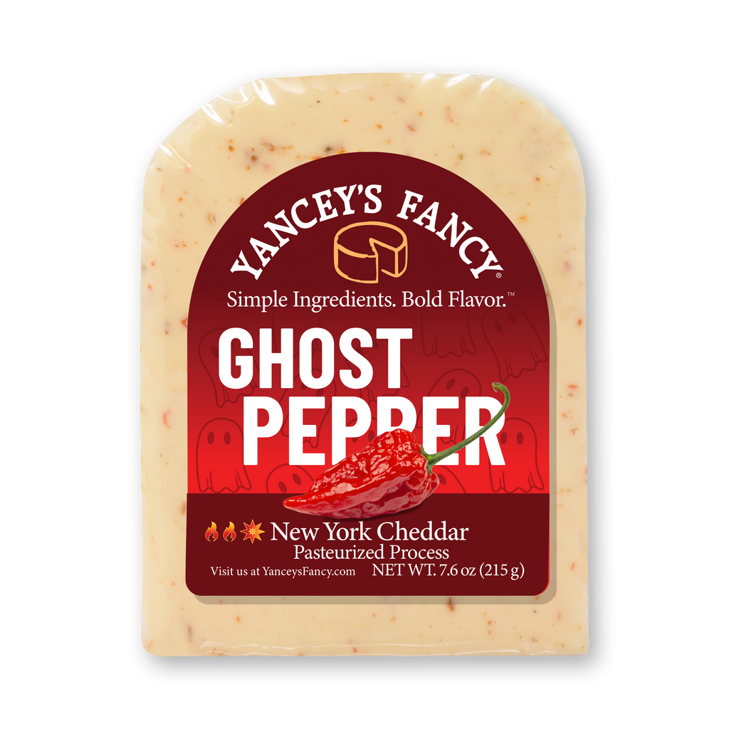 A vacuum-sealed block of Yancey's Fancy "Ghost Pepper" New York Cheddar cheese. The label is primarily deep red with a white arched border, featuring a graphic of a bright red ghost pepper in the center and faint, playful cartoon ghosts in the background. The text reads "Yancey's Fancy" in an arch at the top, followed by the tagline "Simple Ingredients. Bold Flavor." The product name "GHOST PEPPER" is written in large, bold white capital letters. The bottom of the label indicates it is a "New York Cheddar Pasteurized Process" cheese with a net weight of 7.6 oz (215 g), accompanied by small flame and sun emojis to denote heat. Flakes of red pepper are visible throughout the creamy, off-white cheese block.