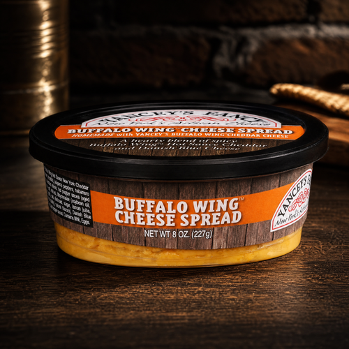 A packaging shot of an 8-ounce (227g) tub of Yancey's Fancy Buffalo Wing Cheese Spread sitting on a dark wood table. The container features a black lid with an orange label, and a matching front label with bold white text reading "BUFFALO WING CHEESE SPREAD" on an orange and faux woodgrain background. The vibrant orange cheese dip is visible through the bottom of the clear tub.