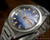 Close-up view of the Osman-Clipper LH-1004 automatic dial, showing the two-tone blue finish, raised gold-tone hour indexes, red seconds hand and Spanish day-date calendar.