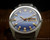 Front view of the Osman-Clipper LH-1004 automatic watch, with two-tone blue dial, raised gold-tone indexes, gold-tone hands, red seconds hand and Spanish day-date display.
