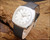 New Old Stock Osman automatic watch displayed on wood, showing the silver sunburst dial, stainless steel case, date display and black leather strap.