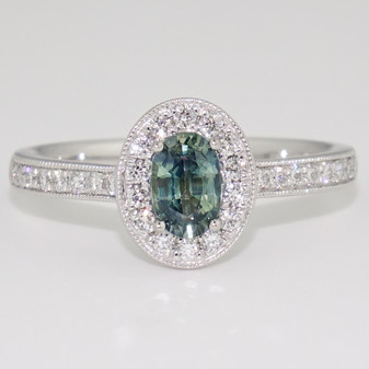 Platinum oval cut teal sapphire and round brilliant cut diamond cluster ring with diamond-set shoulders and a milgrain edge
