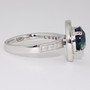 Platinum round cut teal sapphire and diamond halo ring with diamond-set shoulders and milgrain edge side