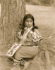 Umatilla girl, full-length portrait, facing front, seated on ground by pine tree, wearing beaded blanket dress, beaded moccasins, holding beaded bag in lap. Poster Print - Item # VARBLL058747572L