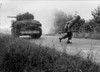 U.S. Soldier Crosses A Road Cautiously Under The Cover Of A Light Tank In The Hedgerows Of Normandy. The Tank Is Equipped With The Hedgecutter Device Developed In July 1944. Normandy Campaign History - Item # VAREVCHISL037EC094