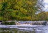 Water cascading over rocks in a flowing river with autumn coloured foliage on the trees; Richmond, North Yorkshire, England Poster Print by John Short (18 x 12)