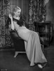 Bette Davis Seated on a Couch with Hand Raised Up in Black Ribbon Top and White Silk Long Dress Photo Print - Item # VARCEL693121