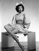 Jane Withers Seated on a Table in White Silk V-Neck Gathered Bodice with Bell Long Sleeve Dress with Knees Bent and Left Hand on the Waist Photo Print - Item # VARCEL705609