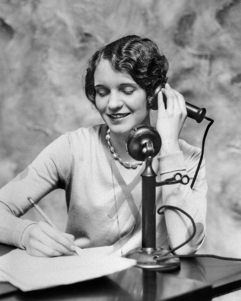 1920s Smiling Woman Sitting At Desk Writing And Talking On