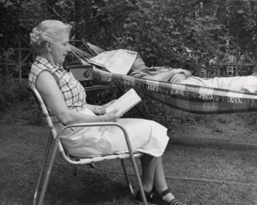 Side profile of a senior woman reading a book with a senior man sleeping in a hammock Poster Print
