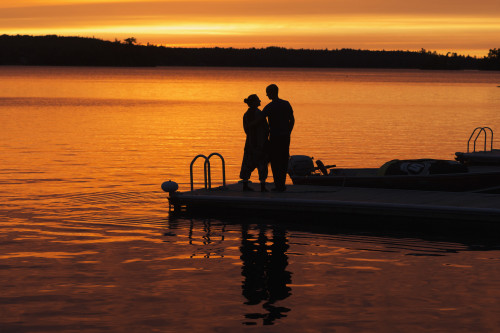 Silhouette of a couple standing at the end of a dock at sunset with an orange glow in the sky and reflected in the tranquil lake water; Ontario, Canada PosterPrint - Item # VARDPI12303007