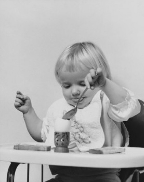Baby sitting in a high chair and eating an egg Poster Print