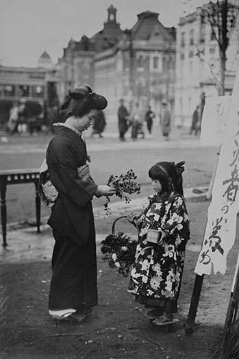 Toddler in front of Kanban Shop sign sells flowers to a kimono wearing young lady who picks them from a basket that the little girls is holding. Poster Print by unknown - Item # VARBLL0587243112