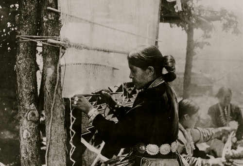 Half-length portrait of Navajo woman, facing left, weaving, and two other Navajo women seated on ground. Poster Print - Item # VARBLL058751311L