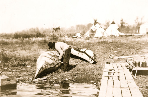 Ojibwa Indian mending canoe at shore and tepees in background. Poster Print - Item # VARBLL058751176L