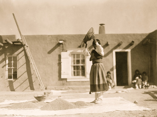 Tewa woman winnowing wheat in front of pueblo building. Poster Print - Item # VARBLL058747590L