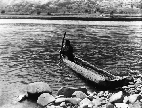 Nez Perc_ Canoe. Nez Perc_ Man With Pole Maneuvering Dugout Canoe To Rocky Shore. Edward S. Curtis History - Item # VAREVCHCDLCGCEC244