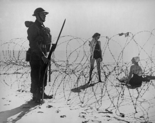 Soldier Standing Guard Next To Barbed Wire Barricade On An English Beach During World War 2. On The Other Side Are Two British Women In Bathing Suits. Sept. 21 History - Item # VAREVCHISL036EC874