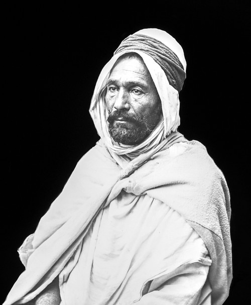 Black and white image of an Arab man against a black background. His outer garment is a 'bournous', a large woollen mantle with a large hood; Algiers, Algeria Poster Print by John Short / Design Pics - Item # VARDPI12516055