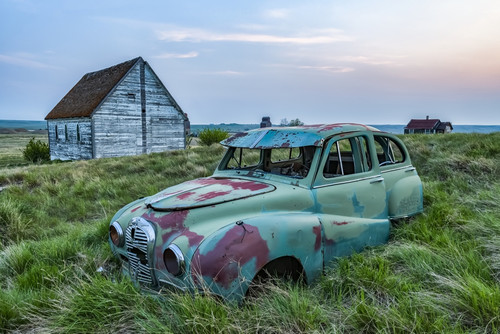 Vintage car sitting in the overgrown grass in a field with old buildings on a farmstead; Saskatchewan, Canada Poster Print by Robert Postma # 12577829
