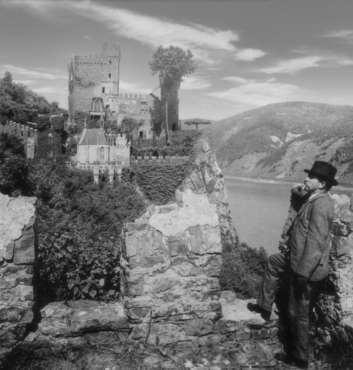 Historic image in black and white of a man standing and smoking a pipe with a view of Rheinstein Castle and the River Rhine, circa 1920; Rhineland-Palatinate, Germany Poster Print by John Short (16 x 17)
