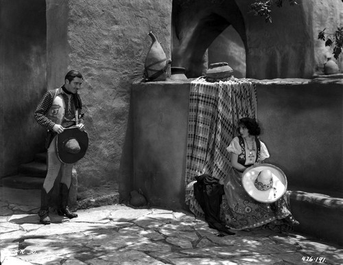 Bebe Daniels Looking at a Man in Cowboy Outfit while Seated Holding a Sombrero in White Blouse and Long Skirt Photo Print - Item # VARCEL696688