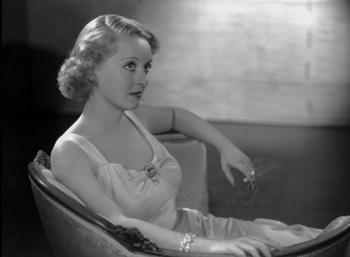 Bette Davis Seated on a Couch with Arms Rested on the Edge of the Chair while Holding a Lighted Cigarette in White Silk Dress Photo Print - Item # VARCEL692796